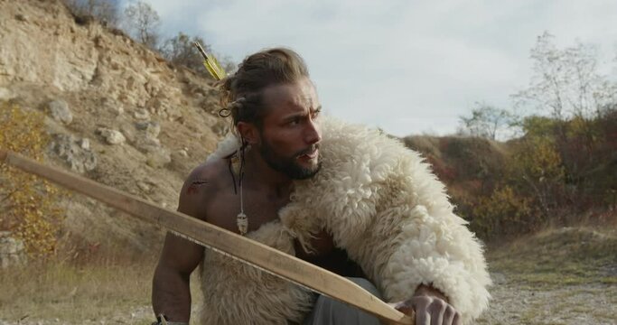 Primitive hunter with bow and arrows clad in a sheepskin tracking his prey in evening light crawling on all fours on the ground - wild caveman
