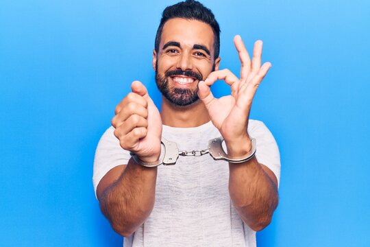 Young hispanic man wearing prisoner handcuffs doing ok sign with fingers, smiling friendly gesturing excellent symbol