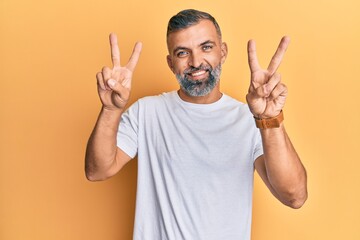 Middle age handsome man wearing casual white tshirt smiling looking to the camera showing fingers doing victory sign. number two.