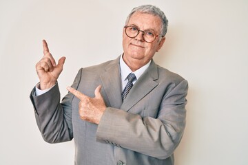 Senior grey-haired man wearing business suit smiling and looking at the camera pointing with two hands and fingers to the side.