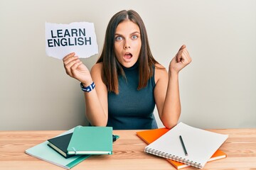 Young caucasian woman holding learn english paper while studying scared and amazed with open mouth for surprise, disbelief face