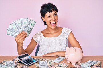 Beautiful brunettte woman holding dollars calculating savings winking looking at the camera with...