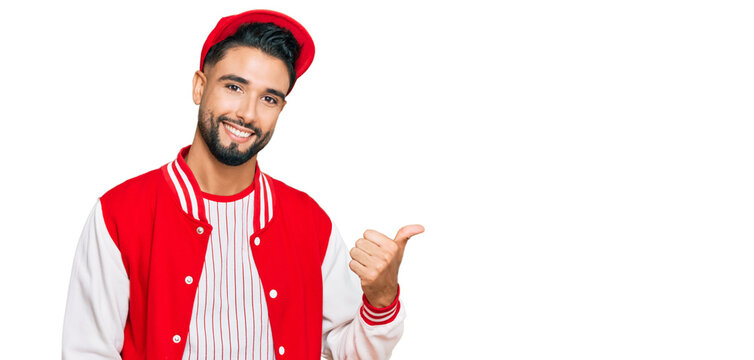 Young man with beard wearing baseball uniform smiling with happy face looking and pointing to the side with thumb up.