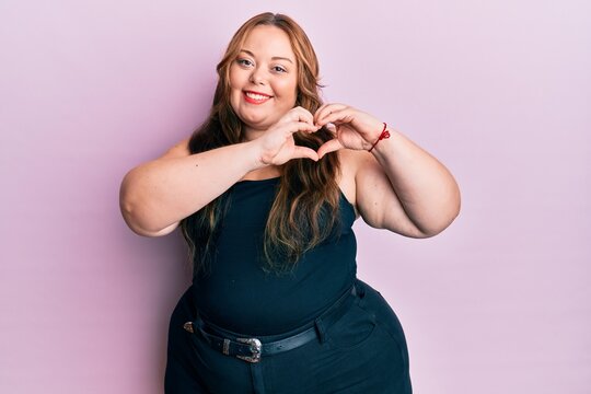 Plus size caucasian young woman wearing casual clothes over pink background smiling in love doing heart symbol shape with hands. romantic concept.