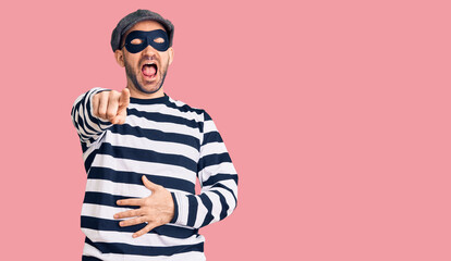 Young handsome man wearing burglar mask laughing at you, pointing finger to the camera with hand over body, shame expression