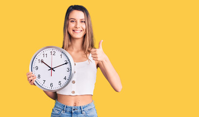 Young beautiful girl holding big clock smiling happy and positive, thumb up doing excellent and approval sign