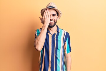 Young handsome man with beard wearing summer hat and shirt covering one eye with hand, confident smile on face and surprise emotion.