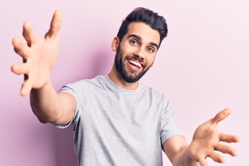 Young handsome man with beard wearing casual t-shirt looking at the camera smiling with open arms for hug. cheerful expression embracing happiness.