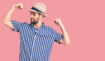 Young handsome man with beard wearing summer hat and striped shirt showing arms muscles smiling proud. fitness concept.