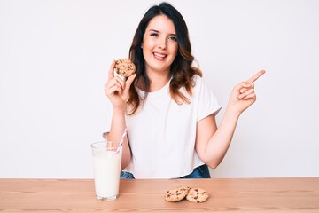 Young beautiful brunette woman drinking a glass of fresh milk with cookies smiling happy pointing with hand and finger to the side