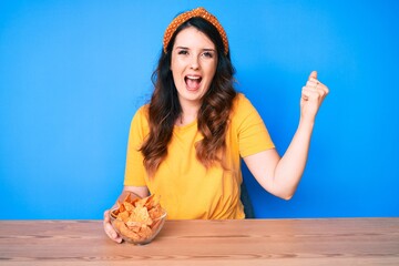Young beautiful brunette woman sitting on the table eating nachos potato chips screaming proud,...