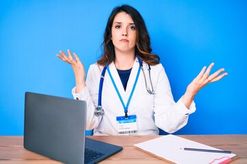 Young beautiful brunette woman wearing doctor uniform working at the clinic clueless and confused...