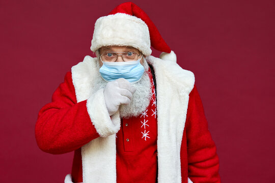 Close up portrait of funny old bearded sick ill Santa Claus wearing costume, glasses, face mask coughing looking at camera standing on Christmas red background. Covid 19 coronavirus concept.