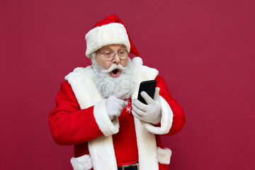 Surprised funny amazed old bearded Santa Claus wearing costume holding cell phone using mobile app...