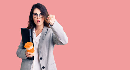 Beautiful young brunette woman holding business folder and drinking coffee annoyed and frustrated shouting with anger, yelling crazy with anger and hand raised