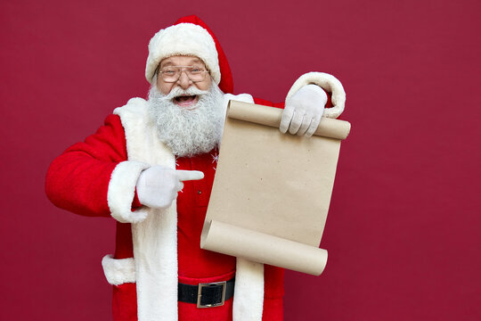 Happy excited old bearded Santa Claus wearing costume holding Merry Christmas wishlist paper roll pointing finger at blank empty xmas wish list letter standing isolated on red background, copy space.