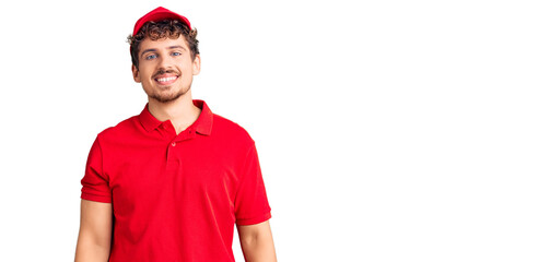 Young handsome man with curly hair wearing delivery uniform looking positive and happy standing and...