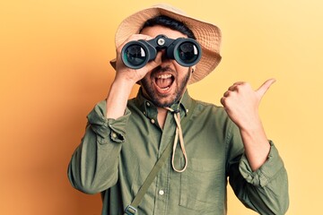 Young hispanic man wearing explorer hat holding binoculars pointing thumb up to the side smiling happy with open mouth