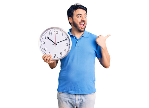 Young hispanic man holding big clock pointing thumb up to the side smiling happy with open mouth