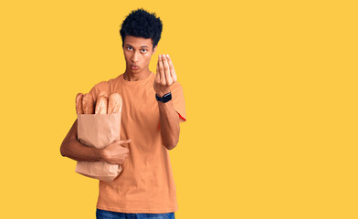 Fototapeta premium Young african american man holding paper bag with bread doing italian gesture with hand and fingers confident expression