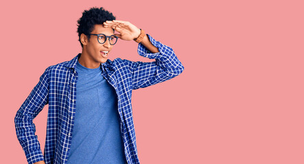 Fototapeta premium Young african american man wearing casual clothes and glasses very happy and smiling looking far away with hand over head. searching concept.