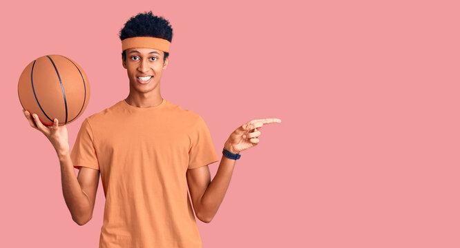 Young african american man holding basketball ball smiling happy pointing with hand and finger to the side