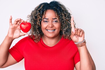 Young african american plus size woman holding heart smiling with an idea or question pointing...