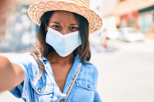 Young beautiful indian woman wearing summer hat and medical mask making selfie by the camera at the city.