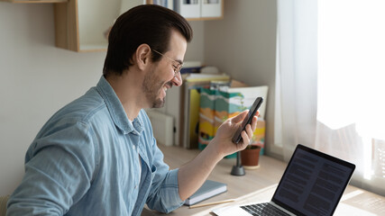 Smiling young man working on computer at home office, reading message on phone with good news, side head shot view. Happy guy looking at mobile screen, involved in pleasant online conversation.