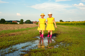 little twin girls in summer yellow dresses and pink boots run through puddles in suburb