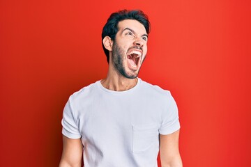 Young hispanic man wearing casual white tshirt angry and mad screaming frustrated and furious, shouting with anger. rage and aggressive concept.