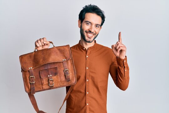 Young hispanic man wearing leather bag surprised with an idea or question pointing finger with happy face, number one