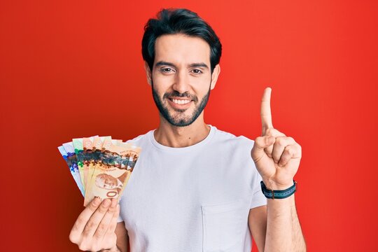 Young Hispanic Man Holding Canadian Dollars Smiling With An Idea Or Question Pointing Finger With Happy Face, Number One