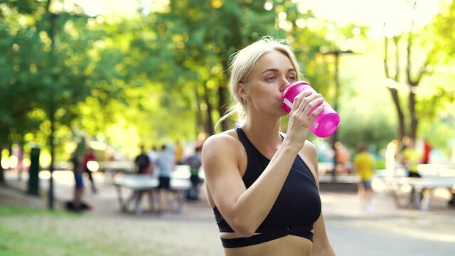 Blonde woman wearing sports top drinking water from plastic bottle while walking in park, blurred people on background. Athletic female after hard training. Concept of fitness