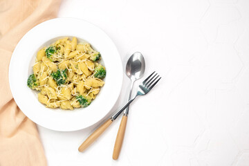 Pasta with green vegetables, broccoli, melted cheese and creamy sauce in white bowl on white table.