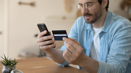Close up focus on plastic credit banking card in male hands. focused young guy using payment information, buying goods by smartphone, happy client purchasing services in mobile application indoors.