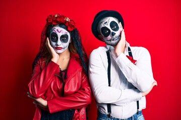 Couple wearing day of the dead costume over red thinking looking tired and bored with depression problems with crossed arms.