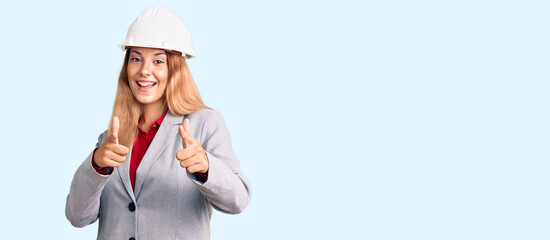 Beautiful young woman wearing architect hardhat pointing fingers to camera with happy and funny face. good energy and vibes.