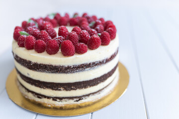 Whole chocolate naked cake with white cheese cream decorated fresh raspberries on a white wooden table, close up.