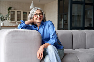 Happy relaxed mature old adult woman wearing glasses resting sitting on couch at home. Smiling mid age grey-haired elegant senior lady relaxing on comfortable sofa looking at camera. Portrait - obrazy, fototapety, plakaty