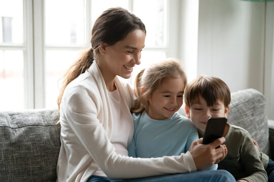Smiling young mom and small kids have fun watching funny video on cellphone gadget together. Happy Caucasian mom relax at home on weekend with little children, use smartphone for entertainment.