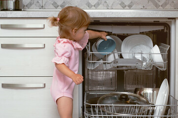 Adorable blonde toddler girl helping in the kitchen taking plates out of dish washing machine
