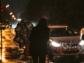 Night city street during the rain. Silhouette of a woman at night in the light of headlights of cars. The woman goes to the taxi car and folds the umbrella. Raindrops flow down from the umbrella.