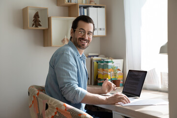 Portrait of happy millennial handsome man in eyeglasses working on computer at home office. Smiling...
