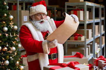Shocked surprised old funny Santa Claus wearing costume holding parchment roll reading letter wish...