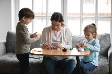 Caring young mom sit at desk with two little children play with plasticine modeling clay together. Happy loving mother or nanny have fun involved in creative activity with playdough with small kids.