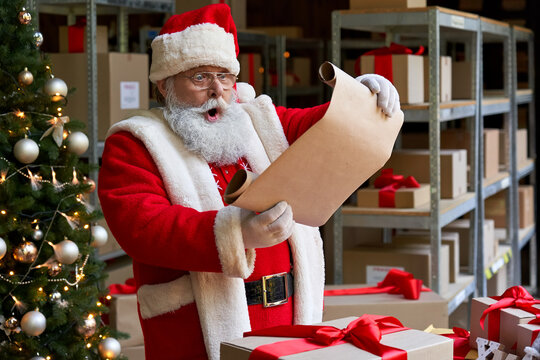 Shocked surprised old funny Santa Claus wearing costume holding parchment roll reading letter wish list preparing Christmas shipping delivery gifts presents parcels standing in workshop on xmas eve.