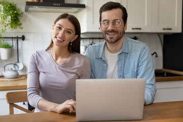 Portrait of happy young loving couple sitting at kitchen table with laptop. Smiling modern technology users clients satisfied with online services, spending leisure time web surfing information.
