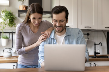 Happy young loving family couple looking at laptop screen, reading e-mail with good news. Smiling handsome man showing funny video on computer to pretty joyful wife, sitting at table in kitchen.