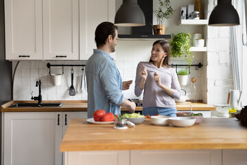 Playful young married couple dancing to energetic music in modern kitchen, distracted from preparing healthy food in modern kitchen. Happy millennial family spouses having fun, enjoying weekend time.